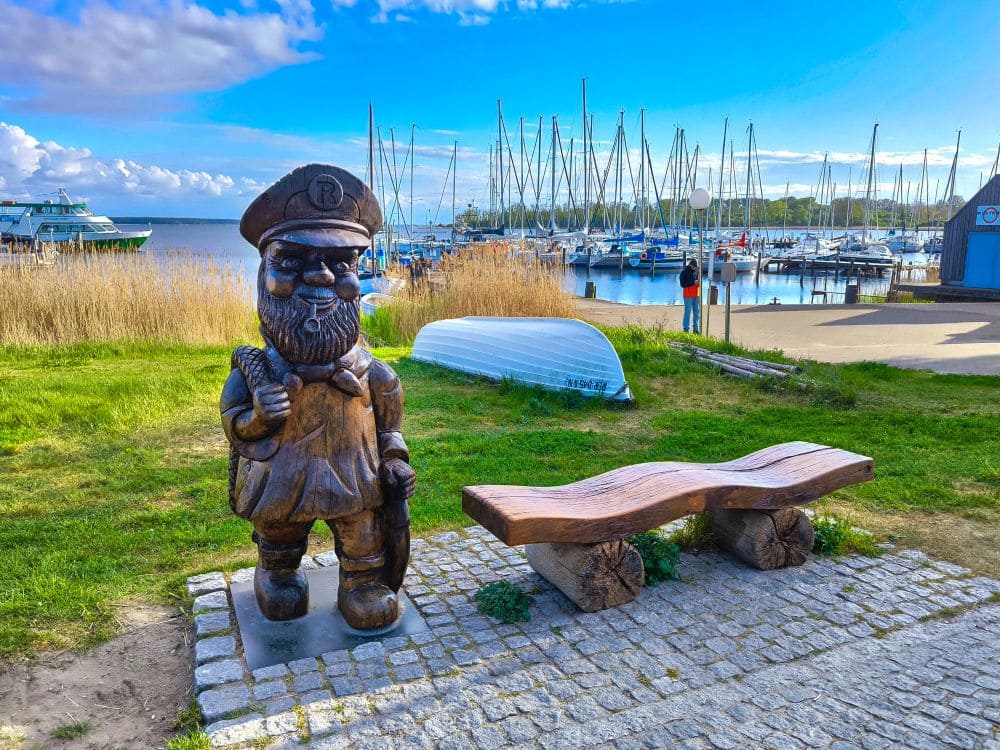 Eine hölzerne Zwergenstatue mit Hut steht neben einer rustikalen Bank auf einem Kopfsteinpflasterweg in der Nähe des Yachthafens im Ostseebad Rerik, wo angedockte Boote, Grasflächen und blauer Himmel die Kulisse für einen idyllischen Urlaub in Rerik bilden.