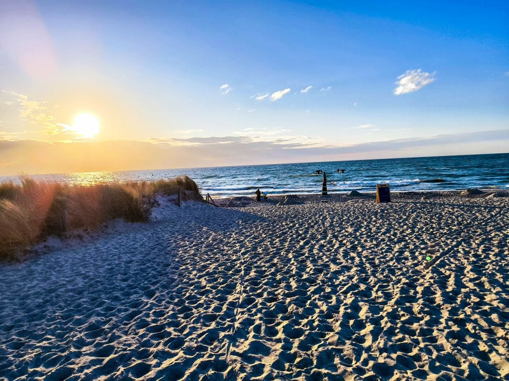 Ein Sandstrand im Ostseebad Rerik bei Sonnenuntergang. Links hohes Gras, sanfte Wellen des Salzhaffs und eine strahlende Sonne tief am Himmel. Am Wasser sind einige Menschen zu sehen, die ihren Urlaub in Rerik und die atemberaubende Aussicht genießen.