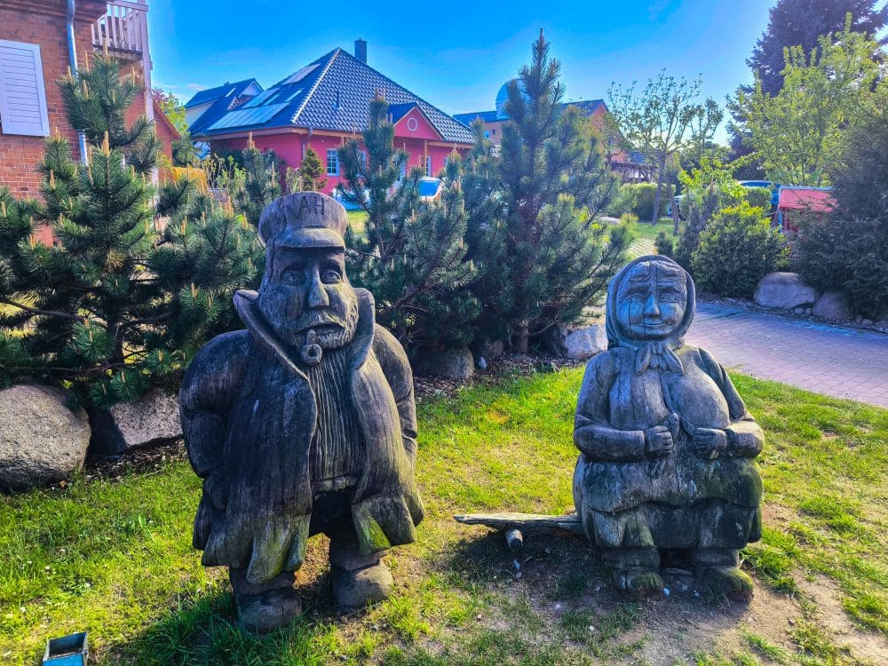 Zwei Holzskulpturen eines älteren Mannes und einer älteren Frau stehen auf Gras in der Nähe von Kiefern in einem Wohngebiet des Ostseebads Rerik, mit roten Backsteinhäusern im Hintergrund, unter einem klaren blauen Himmel – perfekt für einen ruhigen Urlaub in Rerik in der Nähe des Salzhaffs.