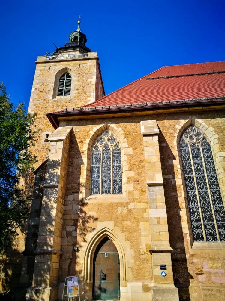 Eine Steinkirche in Kirchheim mit hohen Bogenfenstern, einem Turm und einem roten Schrägdach. Die sonnenbeschienene Fassade steht unter einem klaren blauen Himmel, links ist teilweise ein Baum zu sehen. In der Nähe des Eingangs steht ein Schild.