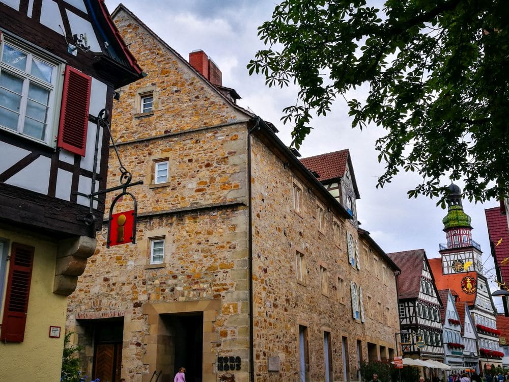 In einer Straßenansicht des malerischen Kirchheims stehen Fachwerkhäuser neben einem Steingebäude. Dekorative Schilder baumeln an den Gebäuden, während sich unter einem leicht bewölkten Himmel Äste über ihnen ausstrecken.