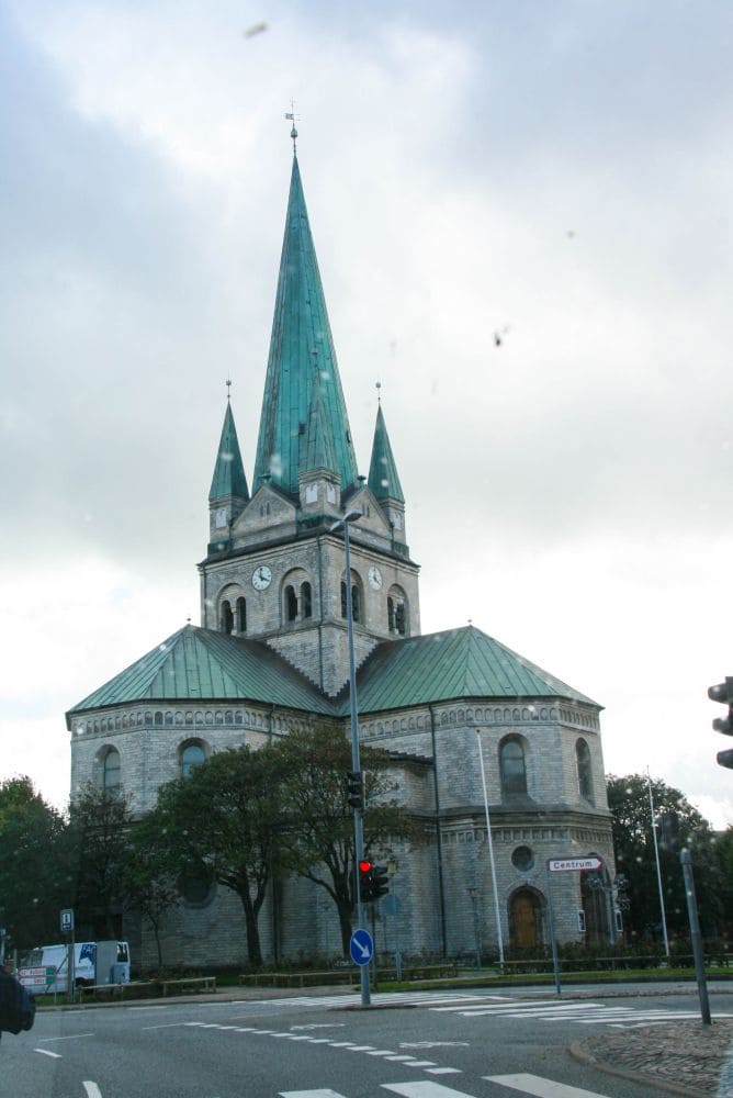 An der Kreuzung steht die große Hurup-Kirche mit ihrem hohen grünen Turm, der von zwei kleineren flankiert wird. Sie besteht aus grauem Stein und steht inmitten von Ampeln und Bäumen, alles unter einem bewölkten Himmel.