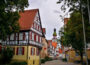 Eine malerische Straße im Dorf Kirchheim besteht aus Fachwerkhäusern mit roten Ziegeldächern. Im Hintergrund ragt ein Kirchturm mit einer Uhr in die Höhe. Die von Bäumen gesäumte Straße liegt unter einem bedeckten Himmel.