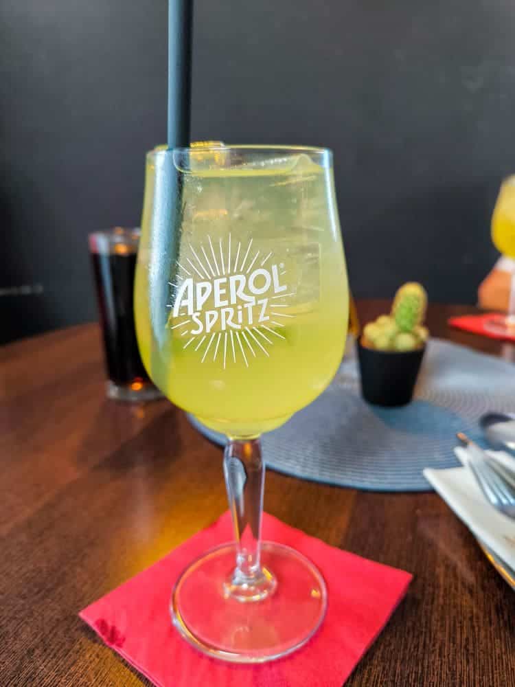 Ein Glas Aperol Spritz mit Strohhalm und Eis steht auf einer roten Serviette auf einem Holztisch und erinnert an die entspannte Atmosphäre von Urlaub in Rerik – eine einladende Szene in einem Ostseebad mit einem Kaktus und Utensilien im Hintergrund.