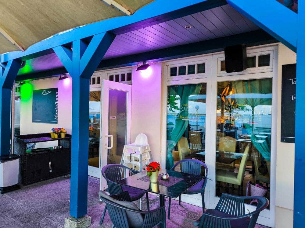 Café-Außenbereich mit schwarzen Tischen und Stühlen, farbenfroher Beleuchtung, einem weißen Hochstuhl und Topfblumen. Glastüren geben den Blick auf die Ostsee und Boote im Hintergrund frei. Blaue Balken rahmen die Terrasse ein – perfekt für Ihren Urlaub in Rerik.