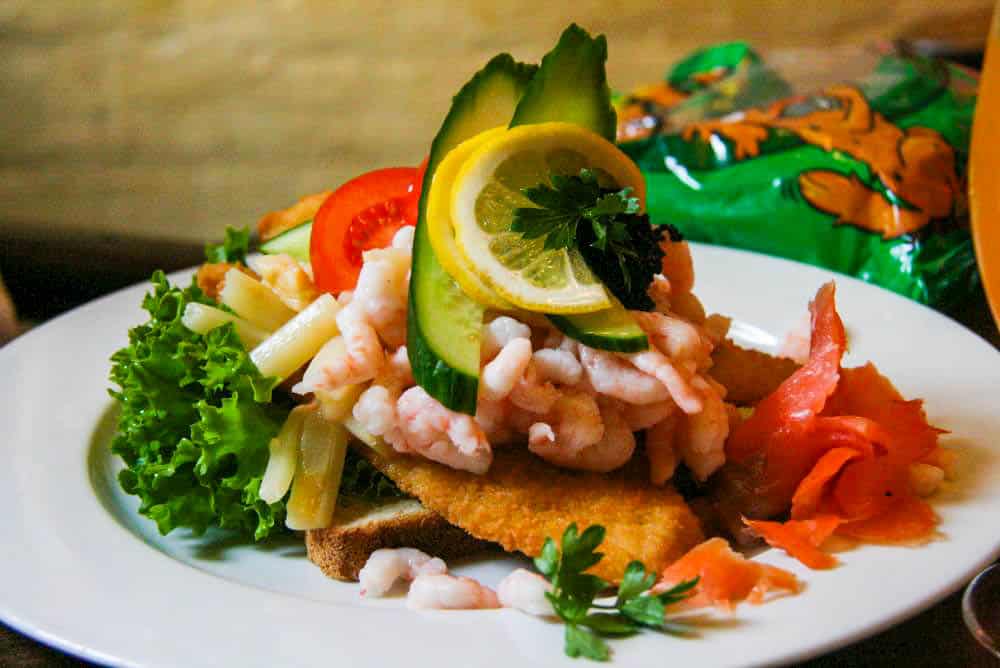 Ein belegtes Hurup-Sandwich besteht aus Garnelen, Zitrone, Gurke, Tomate, Salat und Räucherlachs, elegant angerichtet auf einem panierten Filet. Eine Petersilie als Garnitur krönt die Kreation. Im Hintergrund ist eine Tüte mit Snacks zu sehen.