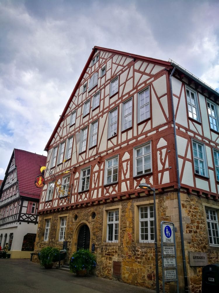 Ein charmantes Fachwerkhaus in Kirchheim verfügt über ein steiles Dach und mehrere Fenster vor einem wolkigen Himmel. Die untere Fassade ist mit aufwendigem Mauerwerk verziert. Ein weiteres ähnliches Gebäude ragt von links hervor, während ein Straßenschild und Topfpflanzen die Vorderseite schmücken.