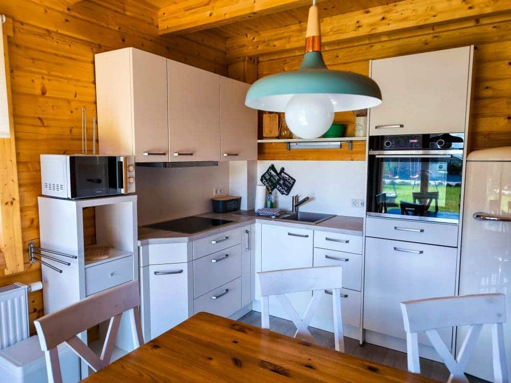 Eine moderne Küche im Ostseebad Rerik mit weißen Schränken, eingebautem Backofen, Mikrowelle, Elektroherd und einem hölzernen Esstisch für vier Personen. Holzwände und -decke sorgen für Wärme, während eine große grün-weiße Pendelleuchte Ihren Urlaub in Rerik abrundet.