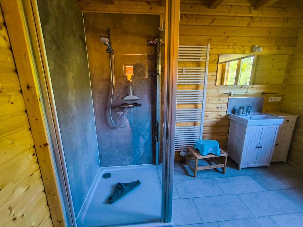 Ein Holzbad mit Glasdusche, weißem Handtuchwärmer, kleinem Waschbecken mit Spiegel und Tritthocker neben einem weißen Waschtisch. Perfekt für einen Urlaub in Rerik am Salzhaff, mit natürlichem Licht, das durch das Fenster strömt. Ein Handtuch liegt auf dem Duschboden.