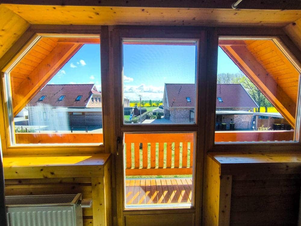 Blick aus einem Holzzimmer durch große Fenster und eine Glastür auf einen Balkon, mit Nachbarhäusern und dem sonnigen, teilweise bewölkten Himmel des Ostseebads Rerik und dem Salzhaff draußen – perfekt für Urlaub in Rerik.