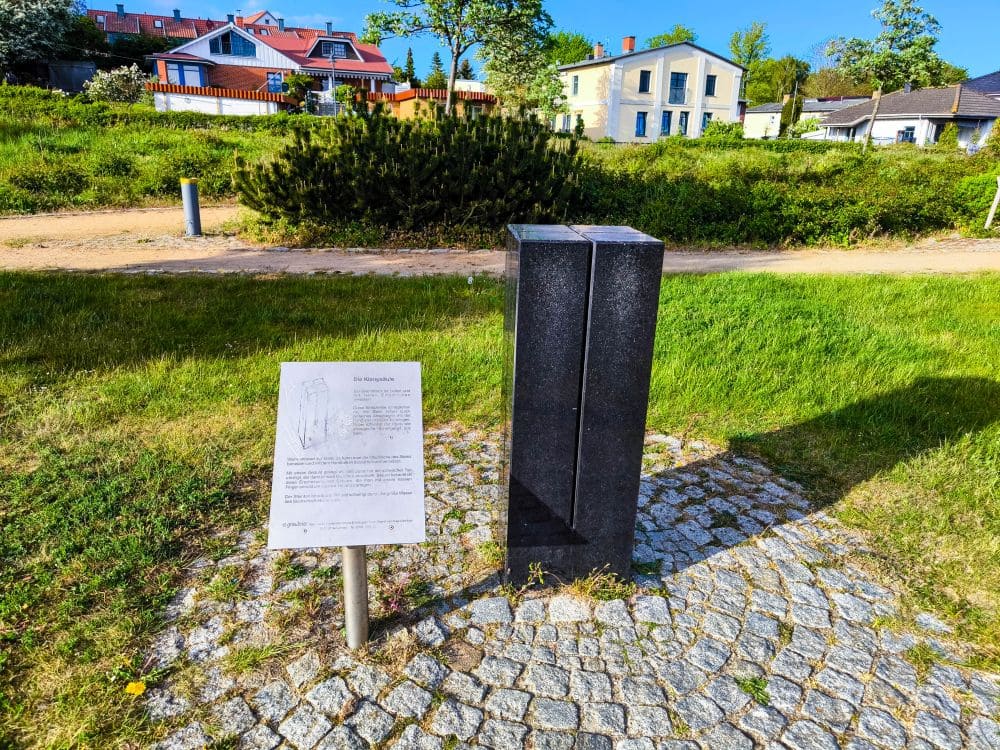 Eine dunkle Steinskulptur steht auf einem Kopfsteinpflasterkreis, daneben ein metallenes Informationsschild. Dieser Ort in der Nähe des Salzhaffs ist perfekt für Ihren Urlaub in Rerik und bietet Rasenflächen, einen Feldweg und Häuser mit roten Dächern unter blauem Himmel.