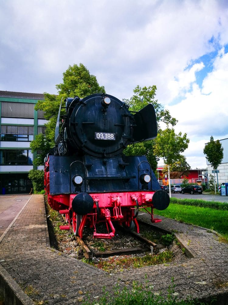 Eine alte schwarz-rote Dampflokomotive steht auf einem kurzen Streckenabschnitt im Freien in Kirchheim. Sie ist von Gras und ein paar Bäumen umgeben, im Hintergrund sieht man ein modernes Gebäude und einen wolkigen Himmel.