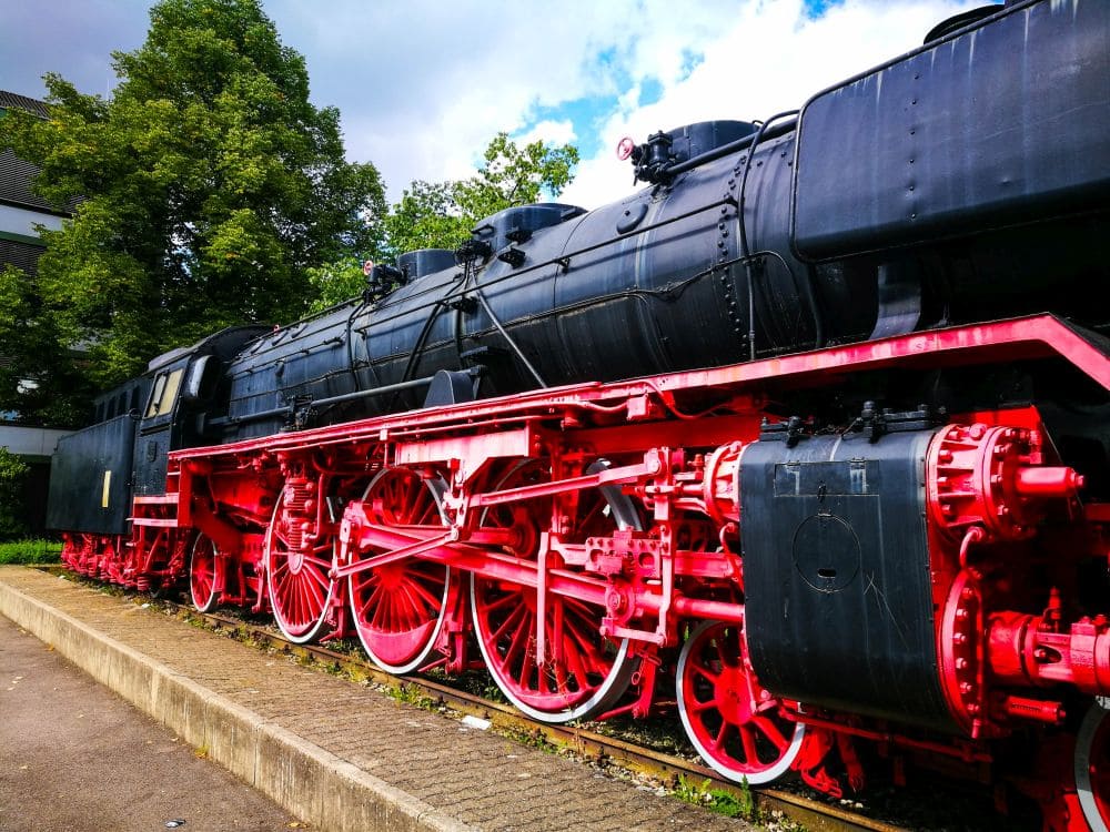 Eine alte Dampflokomotive mit schwarzem Gehäuse und leuchtend roten Rädern steht stolz auf einem Gleis in Kirchheim, umgeben von üppigem Grün und vor einem klaren blauen Himmel.