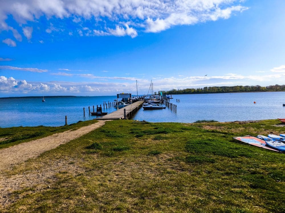 Ein Holzsteg ragt ins ruhige Salzhaff hinein, Boote liegen daneben. Der Himmel ist überwiegend blau mit vereinzelten Wolken, grünem Gras im Vordergrund und Paddleboards am Ufer – eine perfekte Momentaufnahme von Urlaub in Rerik.