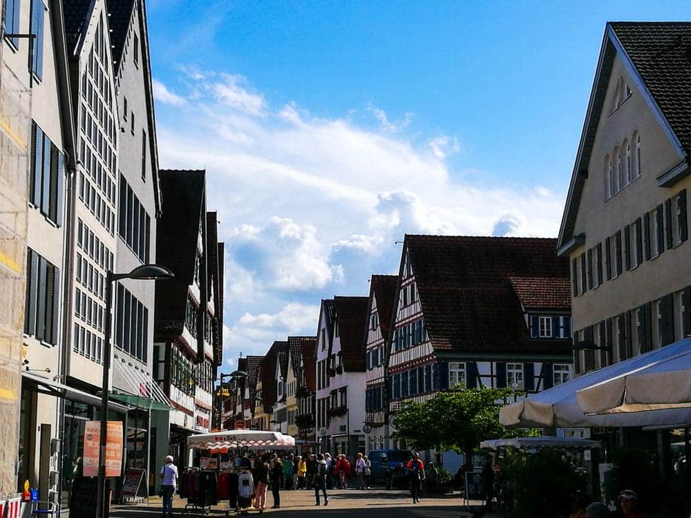 Eine belebte Straße in Kirchheim zeigt traditionelle Fachwerkhäuser unter einem blauen Himmel mit flauschigen Wolken. Menschen schlendern den Kopfsteinpflasterweg entlang und erkunden Marktstände und Cafés mit Sonnenschirmen, die das lebhafte Bild säumen.