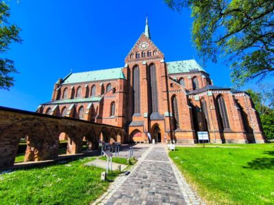 Unter einem strahlend blauen Himmel, umgeben von grünem Gras und Bäumen, steht in Bad Doberan ein großes gotisches Münster aus rotem Backstein. Ein steinerner Weg führt zum Haupteingang, und auf der linken Seite sind gewölbte Backsteinbauten zu sehen.