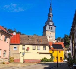Eine ruhige Straße in Bad Frankenhausen, einer kleinen europäischen Stadt, zeichnet sich durch farbenfrohe Häuser und den berühmten Schieferturm aus – einen hohen, schiefen Kirchturm mit Uhr und Turmspitze unter einem strahlend blauen Himmel.