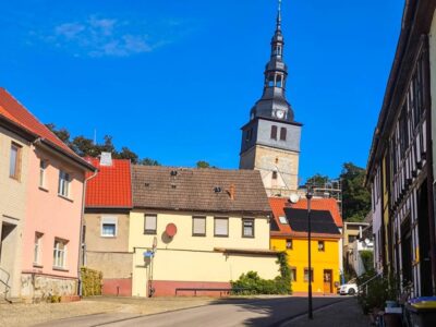 Eine ruhige Straße in Bad Frankenhausen, einer kleinen europäischen Stadt, zeichnet sich durch farbenfrohe Häuser und den berühmten Schieferturm aus – einen hohen, schiefen Kirchturm mit Uhr und Turmspitze unter einem strahlend blauen Himmel.