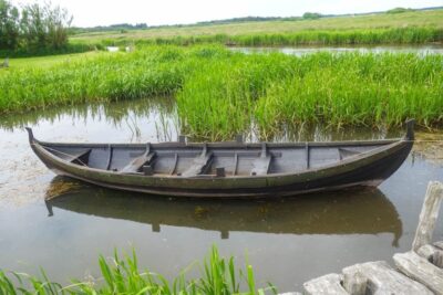 Ein kleines, hölzernes Wikingerboot schwimmt an einem bewölkten Tag auf dem ruhigen Wasser, umgeben von hohem grünem Schilf und grasbewachsenen Feuchtgebieten in der Nähe von Bork Havn - perfekt für eine friedliche Reise.