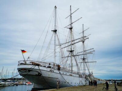 Ein großes weißes Segelschiff mit drei hohen Masten liegt in einem Hafen in einer der historischen Hansestädte vor Anker. Am Heck weht die deutsche Flagge, während mehrere Menschen auf dem kopfsteingepflasterten Steg unter einem bewölkten Himmel stehen.