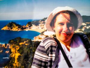 Eine Frau mit weißem Hut und gestreifter Jacke lächelt in die Kamera, während im Hintergrund eine malerische Küstenlandschaft mit blauem Meer und felsigen Hügeln zu sehen ist.