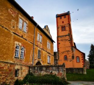 Ein rustikales, orange-gelbes Steingebäude mit weiß umrandeten Fenstern steht neben einem hohen, schmalen Turm in Breitungen. Am wolkenverhangenen Himmel darüber fliegen einige Vögel, und das Grün umgibt die reizvollen Gebäude.