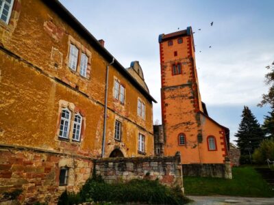 Ein rustikales, orange-gelbes Steingebäude mit weiß umrandeten Fenstern steht neben einem hohen, schmalen Turm in Breitungen. Am wolkenverhangenen Himmel darüber fliegen einige Vögel, und das Grün umgibt die reizvollen Gebäude.