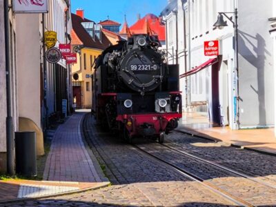 An einem sonnigen Tag fährt eine schwarze Molli-Dampflokomotive eine schmale Kopfsteinpflasterstraße zwischen alten europäischen Gebäuden entlang. Aus ihrer Maschine steigt Dampf auf und an den Ladenfronten sind Schilder zu sehen.
