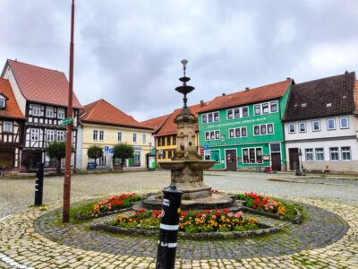 Ein von bunten Blumen umgebener Steinbrunnen steht in der Mitte eines gepflasterten Platzes im idyllischen Städtchen Themar, gesäumt von farbenfrohen Fachwerkhäusern und bemalten Gebäuden unter einem bewölkten Werrataler Himmel.