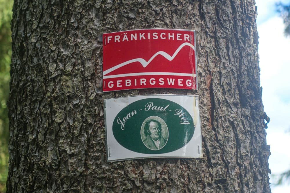 Zwei Wegweiser an einem Baum: Das obere Schild ist rot mit einem weißen Bergumriss und dem Text Fränkischer Gebirgsweg, das untere grün-weiß mit einem Porträt und dem Text Jean-Paul-Weg - beide führen zum malerischen Fichtelsee.