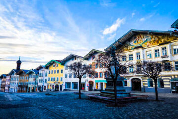 Eine bunte Reihe traditioneller europäischer Gebäude säumt eine Kopfsteinpflasterstraße in Bad Tölz, mit blattlosen Bäumen und einem Brunnen im Vordergrund - eine einladende bayerische Bilderbuchidylle unter einem klaren blauen Himmel, perfekt für Ihre nächste Reise.