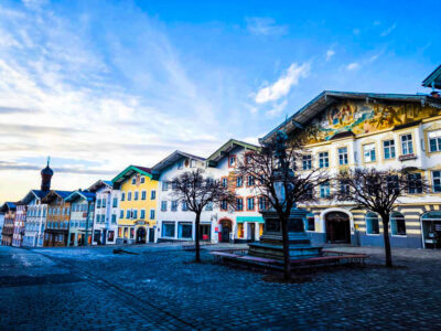Eine bunte Reihe traditioneller europäischer Gebäude säumt eine Kopfsteinpflasterstraße in Bad Tölz, mit blattlosen Bäumen und einem Brunnen im Vordergrund - eine einladende bayerische Bilderbuchidylle unter einem klaren blauen Himmel, perfekt für Ihre nächste Reise.