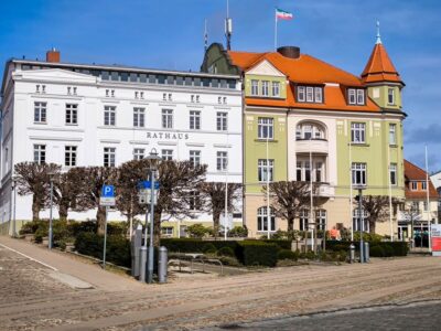 Ein historisches Gebäude mit weißer und grüner Fassade, das als Rathaus bezeichnet wird, steht in einer Kopfsteinpflasterstraße in Bergen auf Rügen - im Herzen der Insel entdecken Sie Rundbogenfenster, ein rotes Ziegeldach, beschnittene Bäume und eine Flagge auf dem Dach.