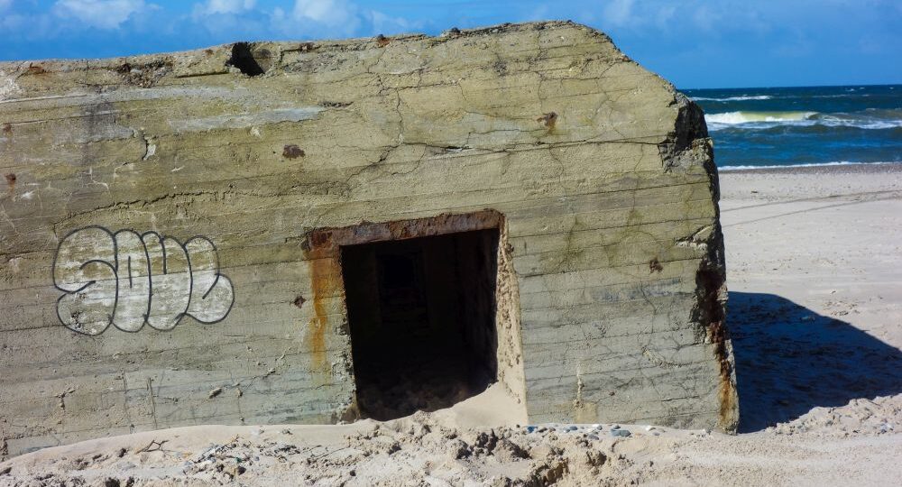 Ein verwitterter Betonbunker mit Graffiti an der Seite steht teilweise im Sand eingegraben an einem Strand an der dänischen Küste, seine Geschichte zeichnet sich gegen das Meer und den blauen Himmel im Hintergrund ab.