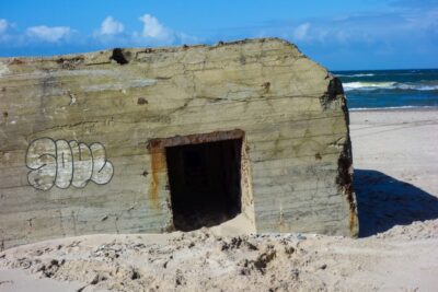 Ein verwitterter Betonbunker mit Graffiti an der Seite steht teilweise im Sand eingegraben an einem Strand an der dänischen Küste, seine Geschichte zeichnet sich gegen das Meer und den blauen Himmel im Hintergrund ab.