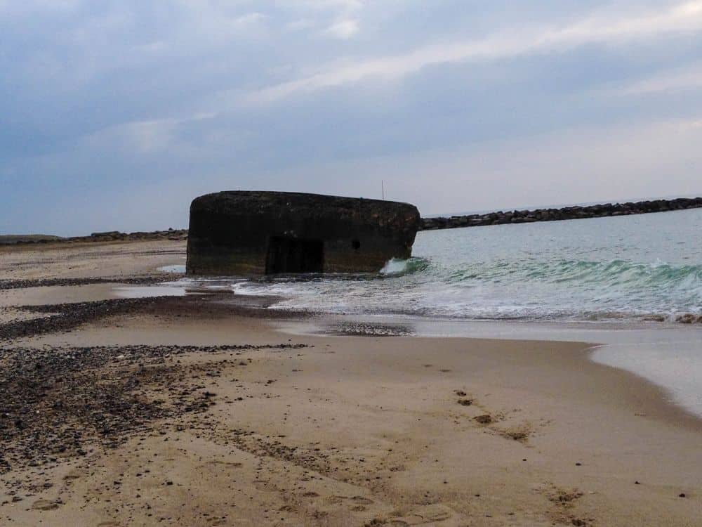 Ein großer Betonbunker, der teilweise im Meerwasser versunken ist, liegt an der dänischen Küste unter einem wolkenverhangenen Himmel, mit sanften Wellen, die ihn umspülen, und einem felsigen Wellenbrecher im Hintergrund, der seine Geschichte andeutet.
