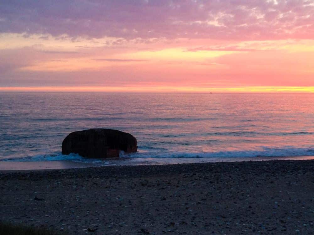 Ein großer Bunker liegt am Rande des Meeres an der dänischen Küste, während sanfte Wellen unter einem leuchtenden, in Rosa, Violett und Orange gefärbten Sonnenuntergangshimmel anrollen. Der Kieselstrand im Vordergrund deutet auf die in die Landschaft geätzten Schichten der Geschichte hin.