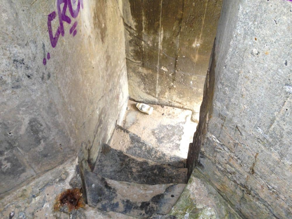 Eine schmale, abgenutzte Steintreppe führt zwischen hohen, verwitterten Betonwänden wie in einem vergessenen Bunker hinab. Eine weggeworfene Tasse liegt am Boden, Graffiti ziert die linke Wand, und ein rostiger Fleck deutet auf eine vergrabene Geschichte hin.