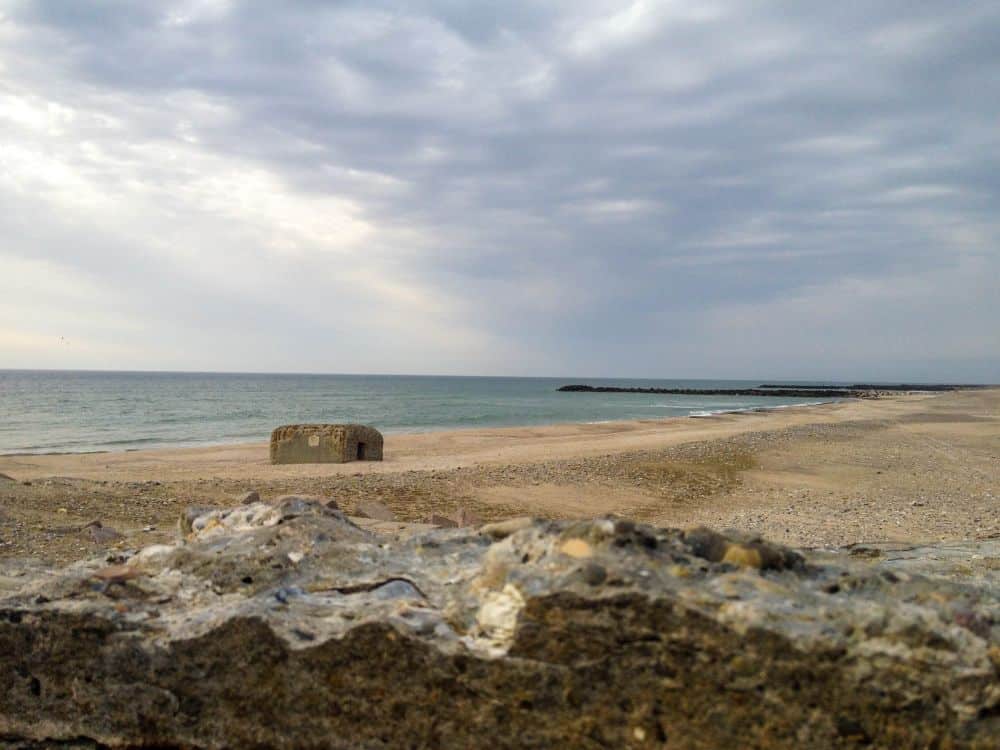 Ein Sandstrand an der dänischen Küste mit einem Betonbunker in der Nähe des Wassers, ruhige Meereswellen und ein bewölkter Himmel. Im Vordergrund deutet eine raue, verwitterte Steinmauer auf die reiche Geschichte der Gegend hin.