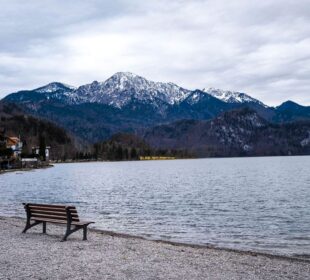 Eine hölzerne Bank steht am ruhigen Kochelsee, mit schneebedeckten Bergen und bewaldeten Hügeln im Hintergrund unter einem wolkenverhangenen Himmel - perfekt für Entschleunigung in der Natur. Auf der linken Seite des Sees stehen Häuser.