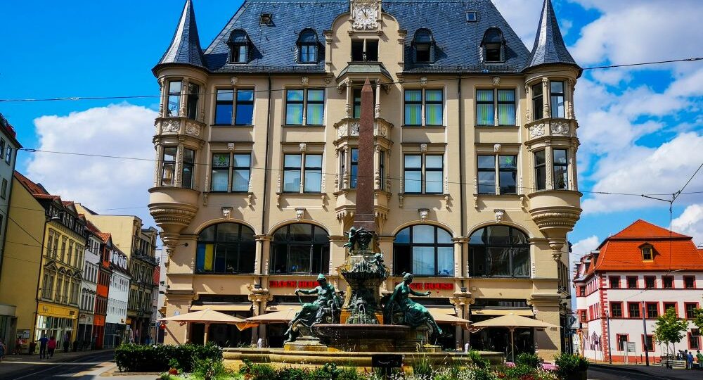 Ein prächtiges historisches Gebäude mit verschnörkelten Türmen und dekorativen Fenstern steht hinter einem großen, kunstvollen Brunnen auf dem sonnigen Stadtplatz von Erfurt, unter einem blauen Himmel mit vereinzelten Wolken.