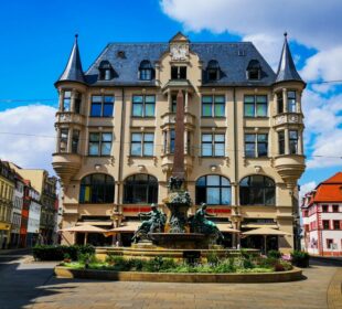 Ein prächtiges historisches Gebäude mit verschnörkelten Türmen und dekorativen Fenstern steht hinter einem großen, kunstvollen Brunnen auf dem sonnigen Stadtplatz von Erfurt, unter einem blauen Himmel mit vereinzelten Wolken.