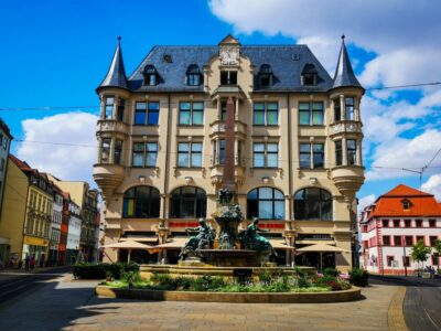 Ein prächtiges historisches Gebäude mit verschnörkelten Türmen und dekorativen Fenstern steht hinter einem großen, kunstvollen Brunnen auf dem sonnigen Stadtplatz von Erfurt, unter einem blauen Himmel mit vereinzelten Wolken.