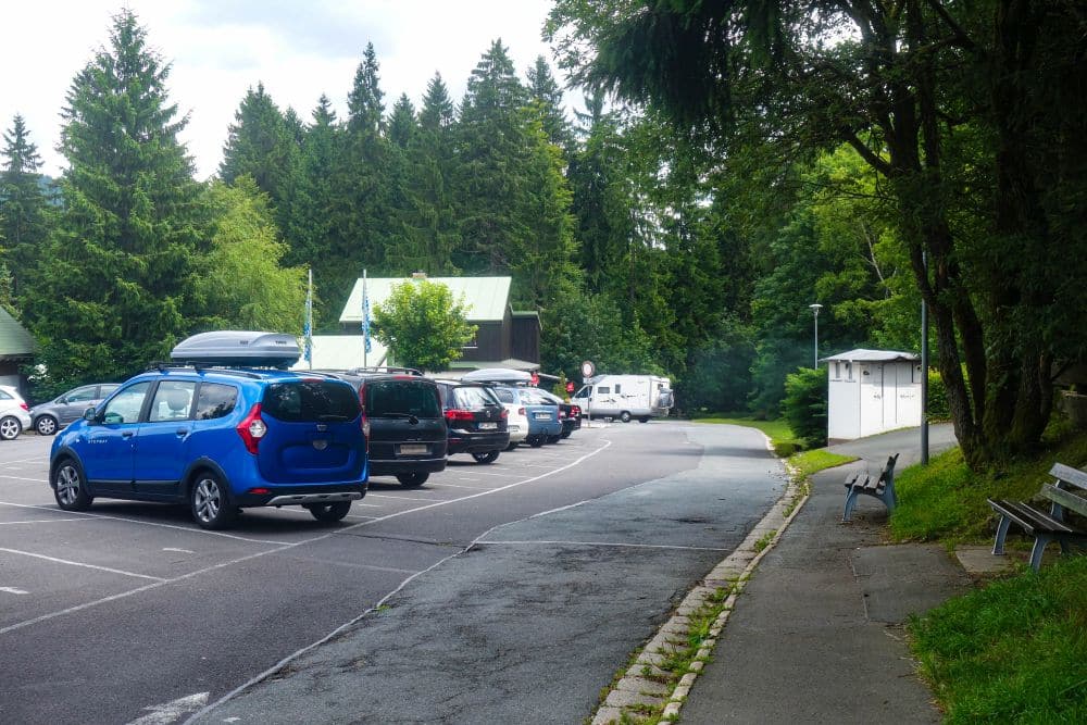 Ein kleiner Parkplatz in der Nähe des Fichtelsees ist von Bäumen umgeben, mit mehreren geparkten Autos, einem weißen Lieferwagen in der Ferne, einem Bürgersteig mit Bänken und einigen kleinen Gebäuden unter einem bewölkten Himmel.