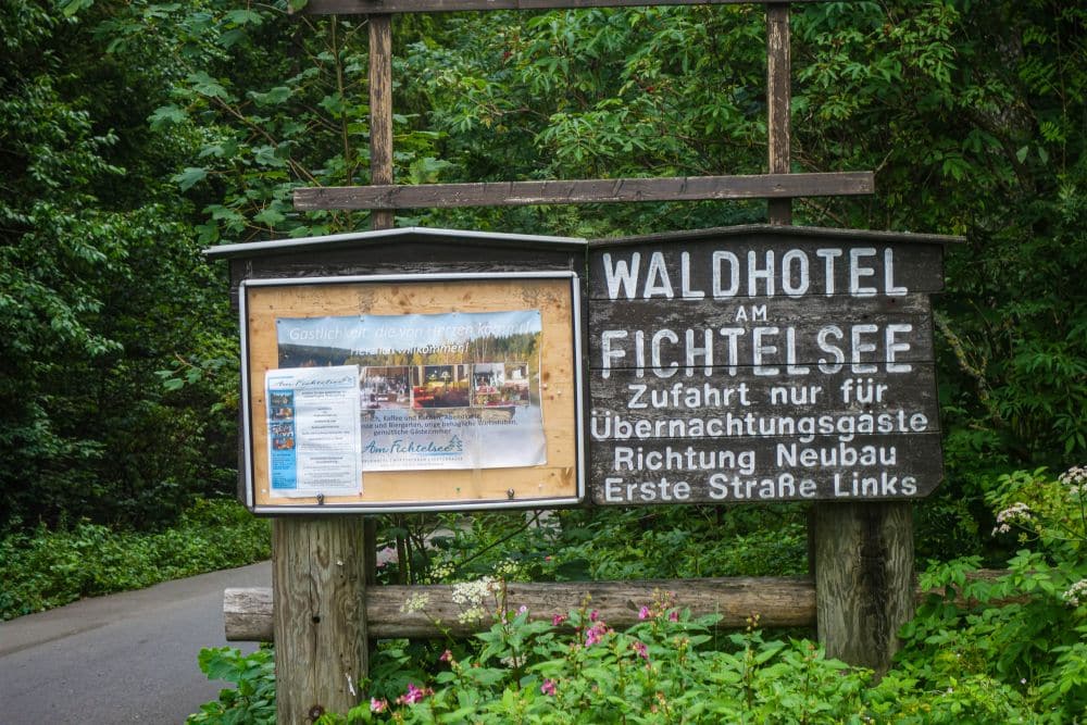 Auf einem Holzschild in einem Waldgebiet in der Nähe des Fichtelsees steht "Waldhotel am Fichtelsee" mit einer Wegbeschreibung in deutscher Sprache. Daneben hängt eine Pinnwand mit Hinweisen und Fotos, alles umgeben von üppigem Grün.