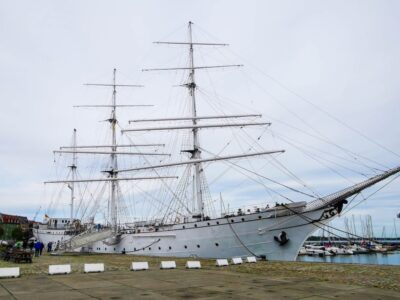 Die Gorch Fock, ein großes weißes Segelschiff mit drei hohen Masten, liegt in einem Hafen vor Anker. Das Schiff ist an einem Steinkai befestigt, seine Takelage und Taue sind sichtbar. Im Hintergrund sind Segelboote und Gebäude unter einem bewölkten Himmel zu sehen.