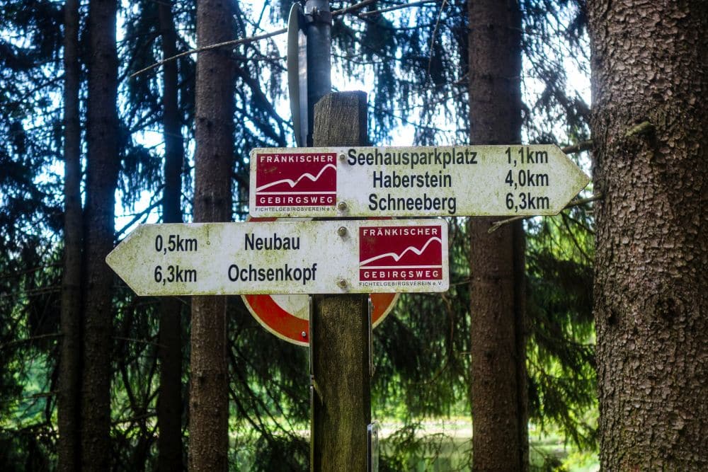 Drei hölzerne Wegweiser in einem Wald weisen auf Ziele wie Seehausparkplatz, Fichtelsee, Haberstein, Ochsenkopf, Neubau und Schneeberg hin, mit Entfernungsangaben in Kilometern. Das Sonnenlicht bricht durch die Bäume im Hintergrund.