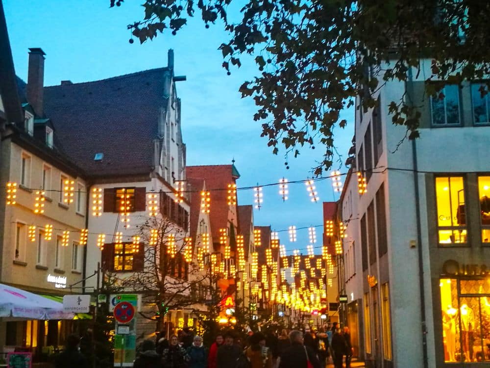 Eine lebhafte Straßenszene in der Abenddämmerung in Ulm zeigt Menschen, die unter Lichterketten zwischen Gebäuden spazieren gehen, wobei Cafés, Geschäfte und Bäume die festliche Atmosphäre noch verstärken. Der blaue Himmel und die belebte Straße fangen die lebendige Energie der Stadt ein.