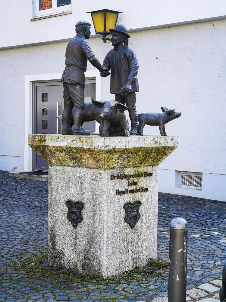 Eine Bronzestatue in Ulm stellt zwei Männer dar, die sich die Hände schütteln, daneben ein Schwein und ein Ferkel. Sie steht auf einem Steinsockel vor einem weißen Gebäude und ist mit einer Inschrift und zwei Ziermasken versehen.