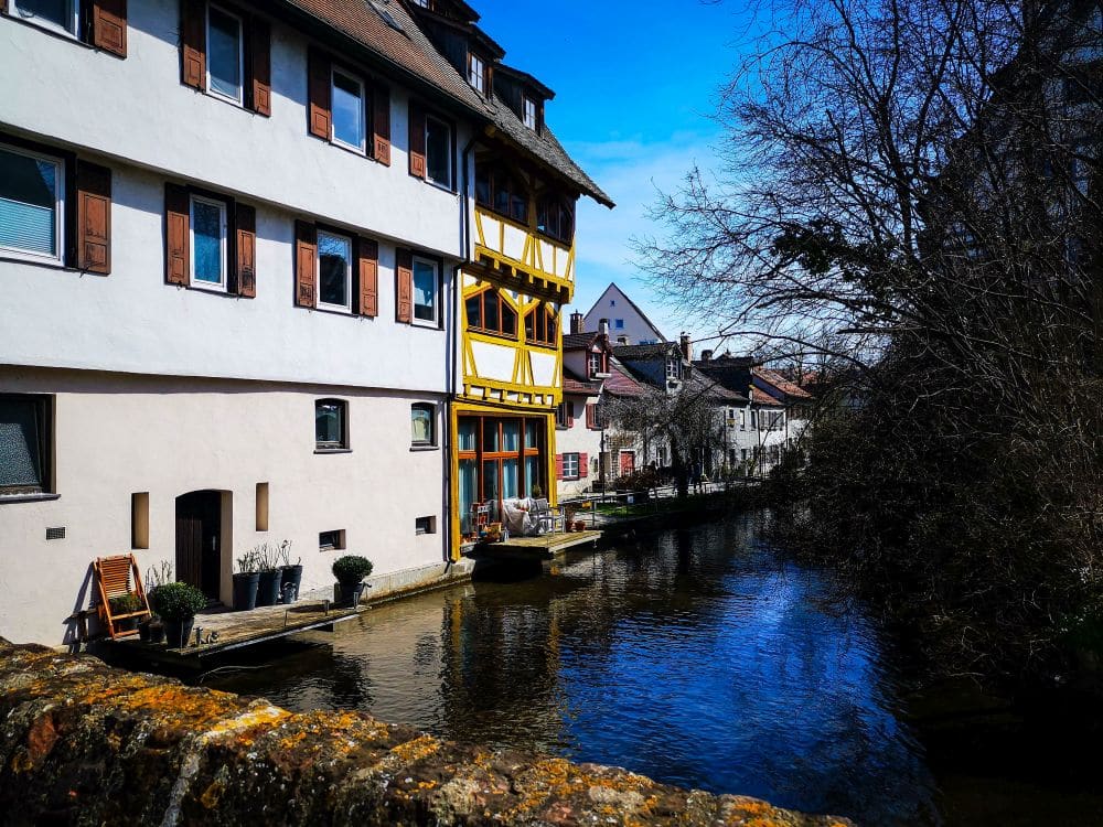 Ein malerischer Kanal in Ulm, gesäumt von alten Gebäuden, darunter ein gelb-weißes Fachwerkhaus, unter einem strahlend blauen Himmel; auf der rechten Seite des Wassers stehen Bäume ohne Blätter.